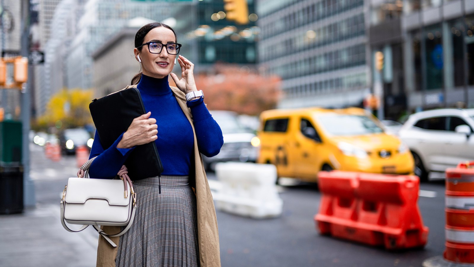 Smiling businesswoman with eyeglasses, wireless headphones and a laptop seen on the street of Manhattan using her wireless headphones after finishing work.