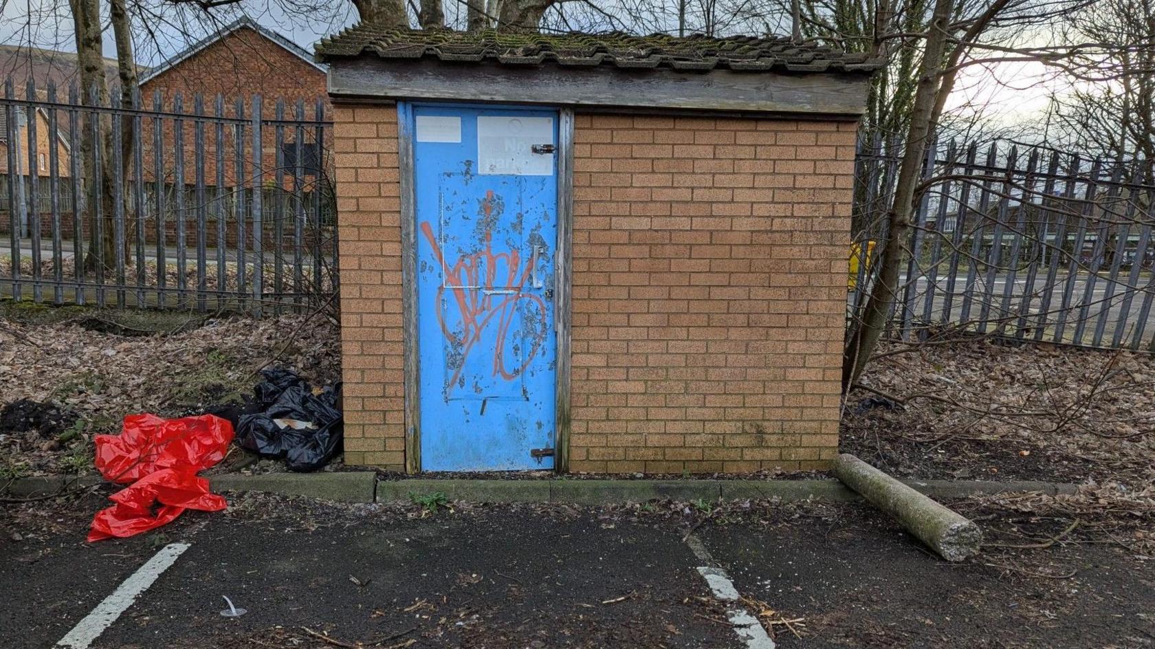 A small brick building with a blue door in a car park. There are plastic rubbish bags strewn beside it.