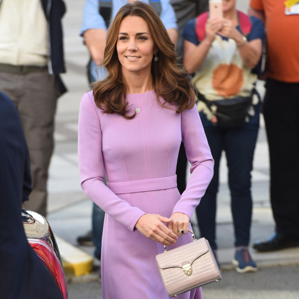  Kate Middleton walking outside with people behind her taking photos while wearing a purple long-sleeved dress holding a lilac top-handled Aspinal bag . 