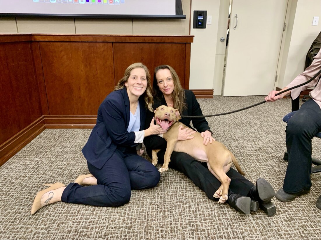 Two smiling women sit on a courtroom floor with a smiling dog.