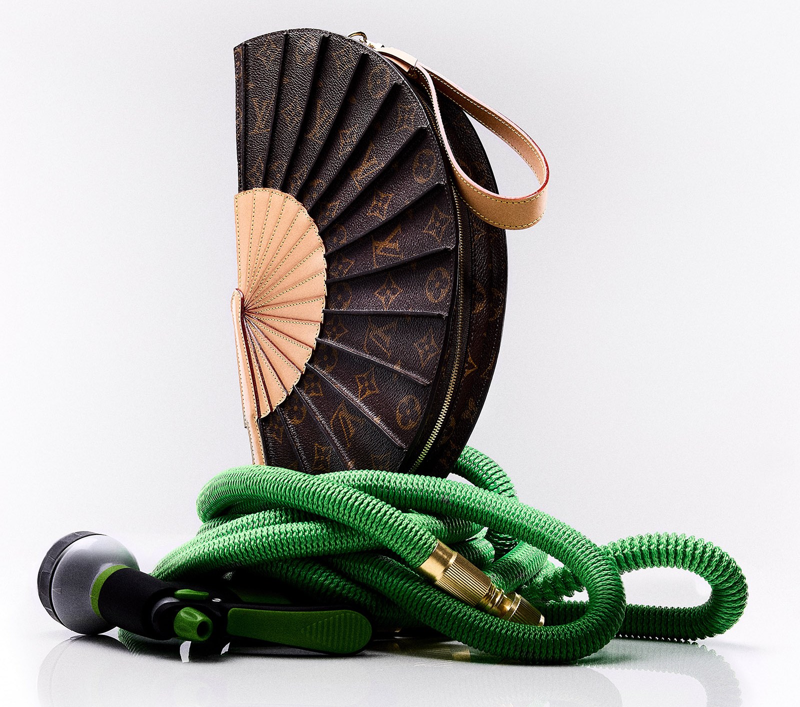 A brown and beige fan-shaped handbag sitting on a coiled green garden hose and nozzle.