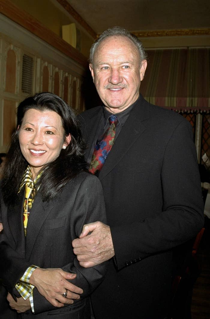 Two people posing together, both in suits. The man has a colorful tie and smiles with his arm around the woman, who also smiles for the photo