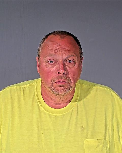 A middle-aged man with short hair and a goatee wearing a yellow T-shirt stands against a plain gray background, looking directly at the camera.